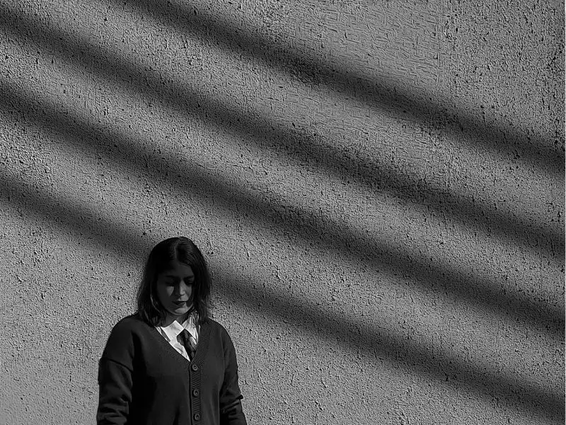 Eine Frau steht im Licht-Schatten-Muster an einer Wand -sinnbildlich für Würde, Verletzlichkeit und gesellschaftliche Vorurteile